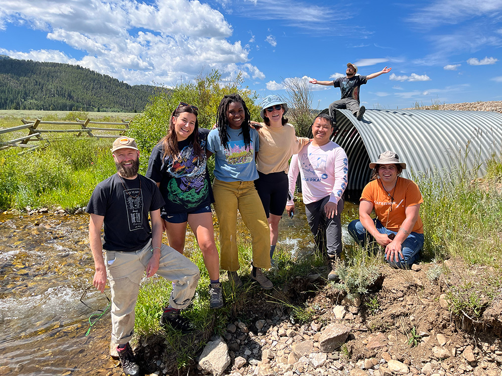 A group of 6 people are outdoors on a sunny day with clear blue skies and a mountain vista in the background. One person is sitting on a metal water tunnel. 